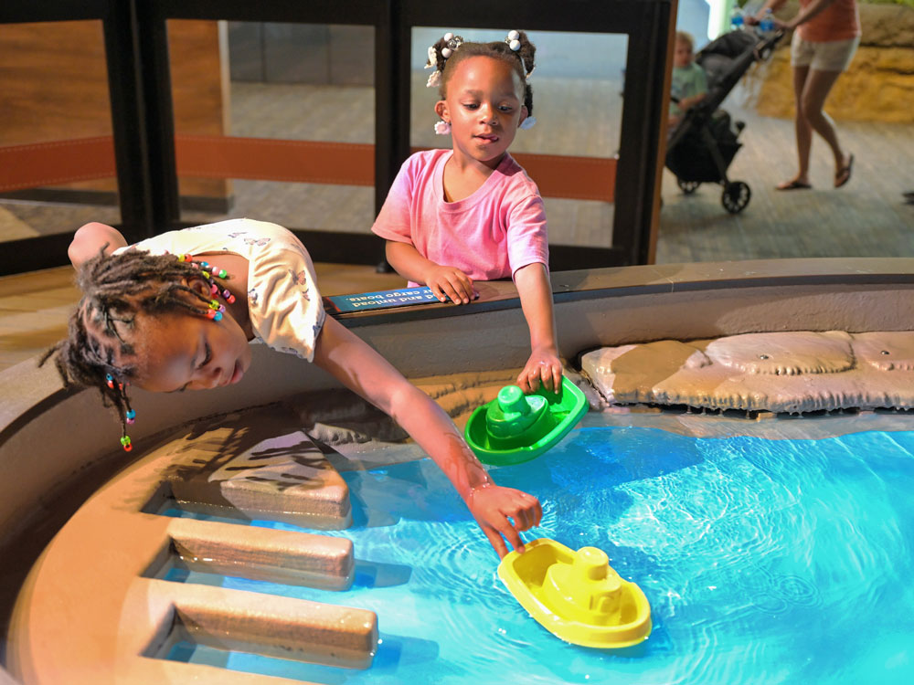 Two children playing with toy boats in an indoor water table.